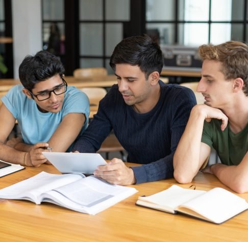 Mentor training students of business school in library. Expert holding tablet and explaining project details, two young men listening to him. Practical training concept